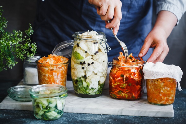 A home chef using a fork to place vegetables into a glass jar for preserving. There are other jars of various sizes and contents on the counter as well.